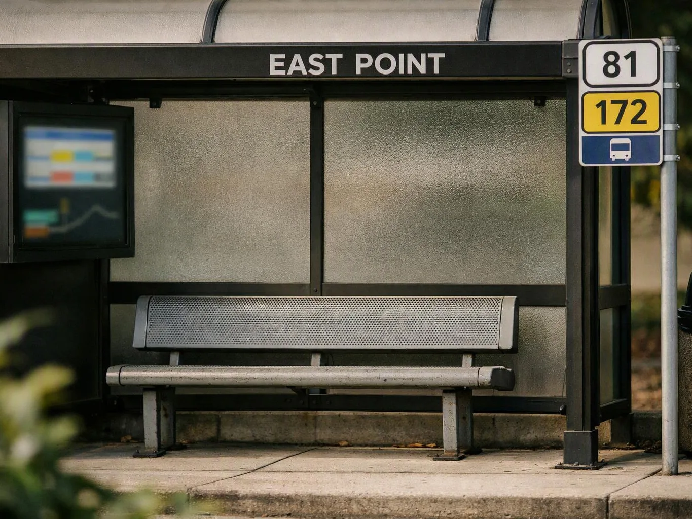 An empty bus shelter with a bench, route signs, and frosted glass panels sits beside a sidewalk